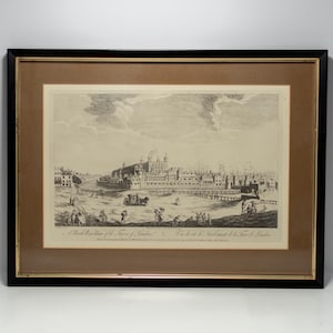 May include: Framed black and gold print of a historical cityscape. The print, featuring a detailed illustration, depicts a city with buildings, a bridge, and figures. The artwork is encased in a black frame with a gold inner border, suitable for home decor.