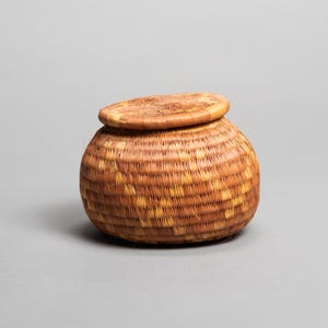 Könnte beinhalten: Ein runder, gewebter Korb mit Deckel. Der Korb besteht aus naturfarbenen Fasern, mit einem Deckel aus dem gleichen Material. Der Korb hat einen Durchmesser von etwa 13 cm und ist 10 cm hoch. Der Deckel liegt oben auf dem Korb.