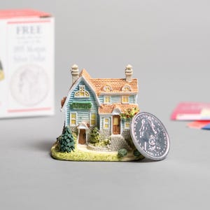 Op de afbeelding: Een miniatuurhuisje met een blauw en groene gevel en een bruin dak. Een zilveren munt ligt naast het huis. Op de achtergrond staat een doos met een afbeelding van een huis.