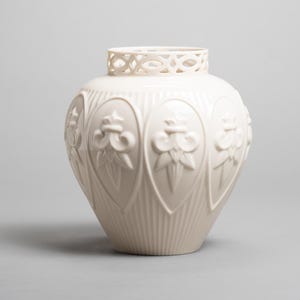 May include: Cream-colored ceramic vase with a round body and a decorative openwork rim. The vase features embossed floral designs within oval frames, and vertical lines on the lower portion. A decorative home decor item.