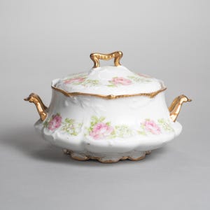 Könnte beinhalten: Eine weiße Zuckerdose aus Porzellan mit Deckel, verziert mit rosa Rosenmustern und goldenen Verzierungen. Die Schale hat zwei goldene Griffe und einen goldenen Griff auf dem Deckel. Der Boden hat einen gewellten Rand.