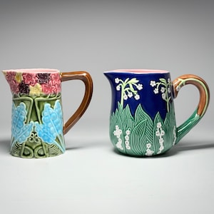 May include: Two ceramic pitchers with floral designs. One pitcher has a blue, green, and pink floral pattern, and the other has a blue background with white flowers and green leaves. Both have brown handles.