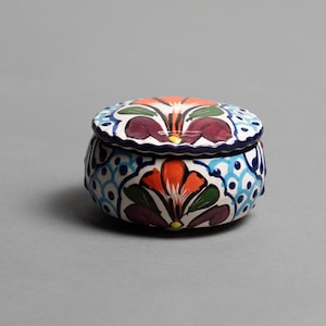 May include: A small, round, ceramic trinket box with a lid. The box is adorned with a colorful floral pattern in shades of red, orange, green, and blue. The lid and base have a dark blue border.