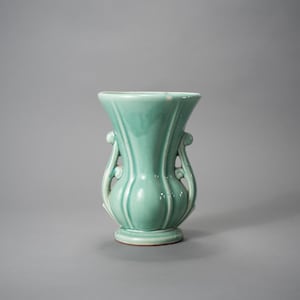 May include: A celadon-colored ceramic vase with a flared top and a rounded base. The vase features vertical ribbed detailing and decorative handles on either side. The vase is approximately 7 inches tall.
