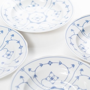 May include: Four white ceramic bowls with a blue floral and vine pattern. The bowls have a scalloped edge and are arranged on a white surface. The blue design includes stylized flowers and connecting vines, creating an elegant aesthetic.