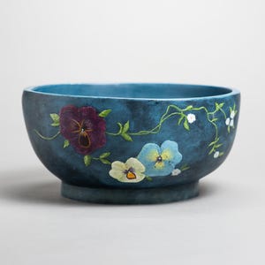 Puede incluir: Un cuenco redondo de cerámica azul con una base azul oscuro. El cuenco está decorado con pensamientos pintados a mano en morado, amarillo y azul, con hojas verdes y flores blancas sobre enredaderas. Es un objeto decorativo.