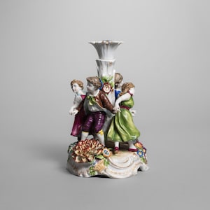 May include: A white porcelain candlestick holder featuring a floral design base and a central column. The candlestick is adorned with figures of children in colorful clothing, holding hands in a circle. The figures are painted in shades of red, green, and brown.