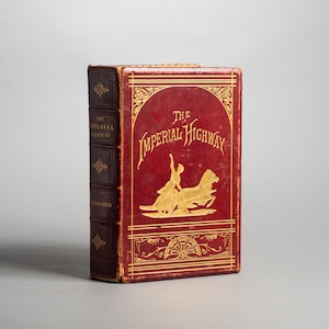 May include: A hardcover book titled The Imperial Highway with a deep red cover and gold embossed lettering. The front cover features a central gold illustration of a person riding a horse-drawn chariot, framed by decorative gold lines and patterns.