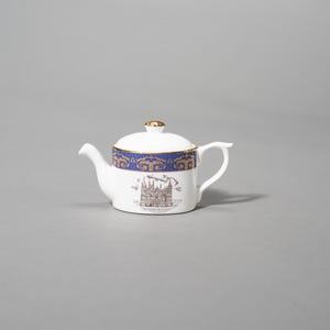 May include: White ceramic teapot with a lid, featuring a blue and gold decorative band around the top and a gold knob. The teapot has a spout and handle, and a brown illustration on the front.