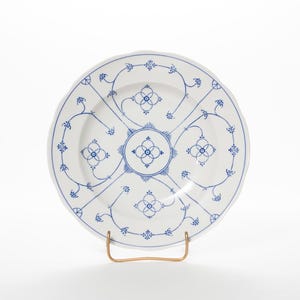 Puede incluir: Plato de cerámica blanca con diseño floral azul. El plato presenta un motivo floral central y un patrón repetido alrededor del borde. El plato se exhibe sobre un soporte de metal dorado.