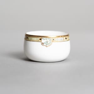 May include: A small white porcelain bowl featuring a gold rim decorated with a repeating arrow pattern and a central shield-shaped emblem containing a light green stylized dragon motif. The bowl has a smooth, rounded base and a clean finish.
