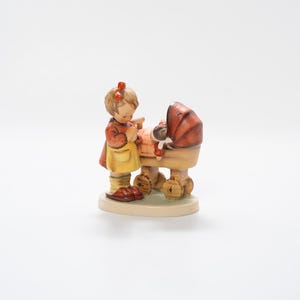 May include: A ceramic figurine of a young girl in a red dress and yellow apron, standing next to a baby carriage. The carriage has a red canopy and a doll inside. The figurine is on a round, off-white base.