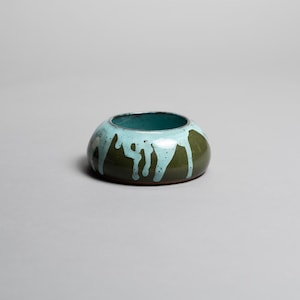 May include: A small, round ceramic bowl with a dark green exterior and a teal interior. The bowl has light blue glaze drips down the sides, creating a unique, artistic look. The bowl is a decorative item.