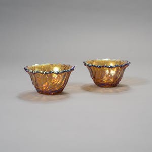 Peut inclure: Deux petits bols en verre orange irisé avec un bord festonné et un motif tourbillonnant texturé. Les bols ont un intérieur doré et un bord bleu-violet foncé. Ils sont probablement décoratifs ou pour le service.