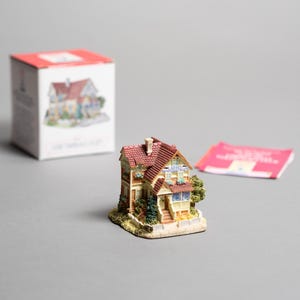 Könnte beinhalten: Eine Miniaturhausfigur mit rotem Dach, beigen Wänden und grünen Sträuchern. Das kleine Haus steht auf einem beigen Sockel. Eine Schachtel mit einem ähnlichen Hausbild und eine rosa Karte befinden sich im Hintergrund.