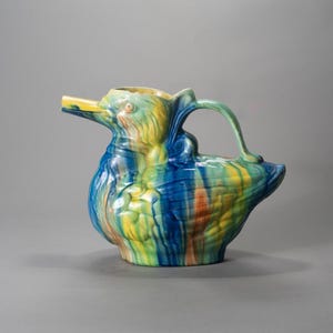 May include: A ceramic pitcher shaped like a duck, featuring a yellow beak and head, with a handle. The body is painted with a blend of blue, green, yellow, and orange hues. The pitcher is a decorative item.