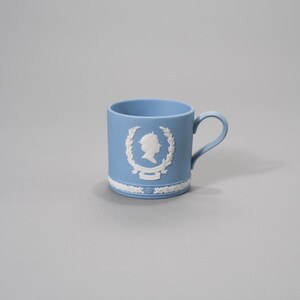 Könnte beinhalten: Hellblaue Keramiktasse mit weißem Reliefdesign. Die Tasse hat einen Henkel und ein dekoratives weißes Relief einer Silhouette in einem Kranz. Der Boden der Tasse hat ein weißes Blumendesign.