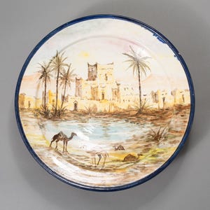 May include: Round ceramic plate with a hand-painted desert scene. The plate features a blue rim and a detailed landscape with palm trees, a fortress, and camels. The color palette includes beige, brown, and blue.