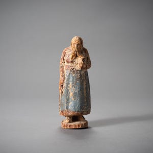 May include: A small, aged wooden figurine of a woman. The figure wears a headscarf, a red top, and a blue skirt. The carving shows signs of wear and tear, suggesting age. The base is a simple, round shape.