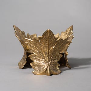 May include: A gold-colored, decorative bowl with a leaf design. The bowl is made of metal and features detailed leaf patterns. The leaves are arranged to form the bowl's sides and base, creating a nature-inspired accent piece.