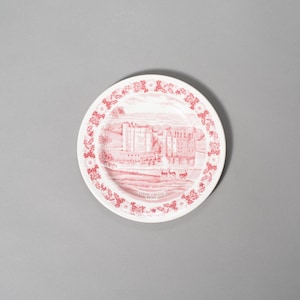 May include: A white ceramic plate with a red transferware design. The plate features a detailed illustration of a castle scene in the center, surrounded by a floral border. The plate is round and has a classic, vintage aesthetic.