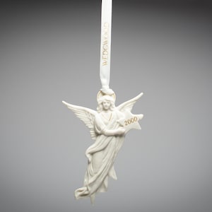 Op de afbeelding: Een wit keramiek engelenornament met uitgespreide vleugels, met een stervormig object met het jaartal "2000". Het ornament hangt aan een wit lint met "WEDGWOOD" erop.