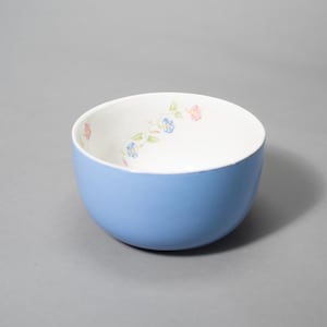 May include: A light blue ceramic bowl with a white interior. The interior has a floral pattern with pink and blue flowers and green leaves. The bowl is round and sits on a grey surface.