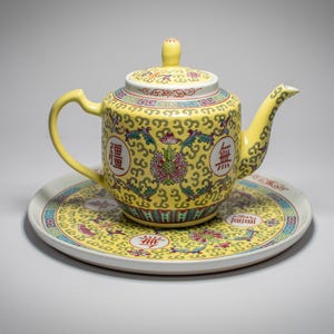 Könnte beinhalten: Eine gelbe Keramik-Teekanne mit passender Untertasse. Die Teekanne und die Untertasse sind mit aufwendigen Blumenmustern und chinesischen Schriftzeichen verziert. Die Teekanne hat einen gebogenen Ausguss und Henkel sowie einen Deckel mit einem kleinen Knopf.