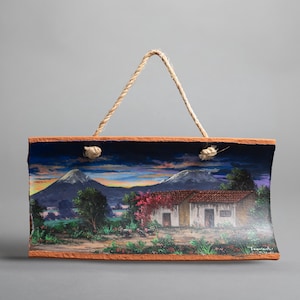 May include: Rectangular wall art featuring a landscape painting of a house with a red tile roof, mountains, and trees. The artwork has a rope hanger and is framed with a brown border. The painting depicts a sunset scene.