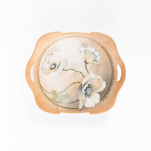 May include: A decorative, antique serving dish with a peach-colored background and a floral design. The dish features white flowers with yellow centers and green stems. The edges have a scalloped design and small handles.