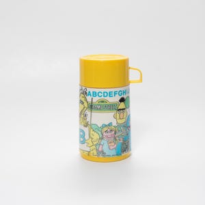 Pode incluir: Uma garrafa térmica amarela da Vila Sésamo com tampa e alça a condizer. A garrafa térmica apresenta personagens de desenhos animados e o alfabeto. A base e a tampa são amarelas, enquanto o corpo é branco com ilustrações coloridas. Um item infantil clássico.