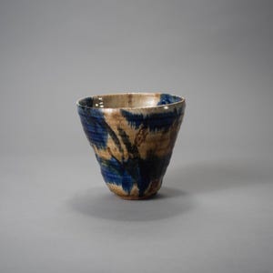 Może przedstawiać: Ręcznie wykonany ceramiczny kubek o unikalnym wzorze. Kubek ma teksturowaną powierzchnię z mieszanką kolorów niebieskiego, beżowego i białego. Wnętrze jest w jaśniejszym odcieniu beżu. Kubek ma lekko rozszerzający się kształt.