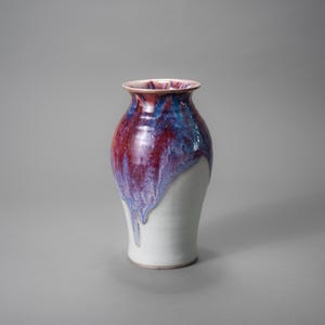May include: A ceramic vase with a white base and a colorful upper section. The upper portion of the vase features a blend of red, blue, and purple hues that drip down the sides. The vase has a wide body and a narrower neck.
