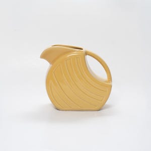 May include: A light yellow ceramic pitcher with a curved handle and spout. The pitcher has a rounded body with a series of curved, vertical ridges. The design is Art Deco inspired.