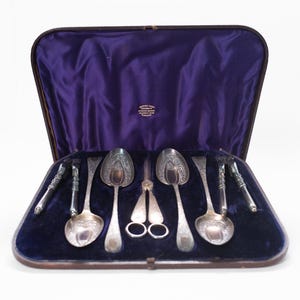 Puede incluir: Un juego de cubiertos de plata antiguos exhibido en un estuche forrado de terciopelo. El juego incluye cucharas, tenedores y utensilios para servir. El estuche es marrón con un interior morado oscuro. Las piezas de plata tienen diseños florales ornamentados.