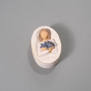 Könnte beinhalten: Eine cremefarbene ovale Schmuckschatulle mit einem geschnitzten Design. Der Deckel zeigt eine Figur, die blaue Blumen hält, mit den Worten "THANK YOU" an der Basis. Die Box befindet sich auf grauem Hintergrund.