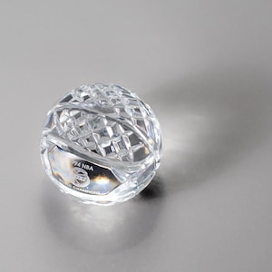 May include: A clear crystal basketball-shaped ornament. The ball features a detailed diamond-cut pattern and a small, oval plaque with the text "94 NBA WORLD CHAMPIONS". The item is on a grey surface.