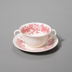 May include: A white teacup with two handles and a matching saucer, both adorned with a red floral pattern. The teacup and saucer are set against a gray background, showcasing a classic tea set design.