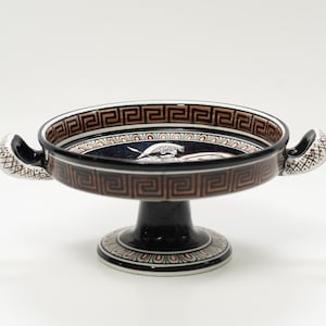May include: A ceramic pedestal bowl with a dark blue interior and a brown and black geometric border. The bowl has two handles and a black base with a decorative pattern. The interior features a white illustration.