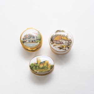 Puede incluir: Tres joyeros decorativos con bordes dorados y pinturas detalladas de paisajes en las tapas. Las cajas presentan imágenes de edificios y paisajes, sobre un fondo blanco.