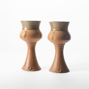 Puede incluir: Dos copas de cerámica hechas a mano. Las copas tienen una base marrón y un borde verde claro. El cuerpo de las copas tiene una forma bulbosa con ranuras verticales. Las copas miden aproximadamente 25 cm de altura.