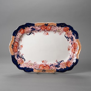 Könnte beinhalten: Eine ovale Servierplatte mit gewelltem Rand. Der Rand ist marineblau mit orangefarbenen und rosa floralen Designs. Die Mitte ist weiß. Die Platte hat einen dekorativen, gemusterten Griff.