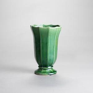 May include: A green ceramic vase with a fluted design. The vase has a wide, scalloped rim and a pedestal base. The color is a rich, deep green, and the surface has a glossy finish. The vase is approximately 8 inches tall.