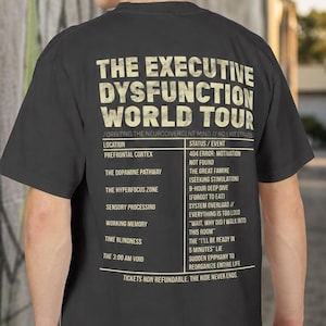 May include: A dark gray short-sleeve t-shirt featuring a beige graphic on the back. The text reads THE EXECUTIVE DYSFUNCTION WORLD TOUR followed by a list of locations and events including PREFRONTAL CORTEX, THE DOPAMINE PATHWAY, and TIME BLINDNESS.