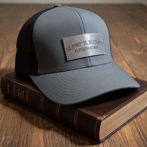 May include: Gray baseball cap with a black mesh back and a silver rectangular patch. The patch displays the coordinates 32.0987° N, 35.2310° E and an elevation of 740m. The cap is resting on a brown book.