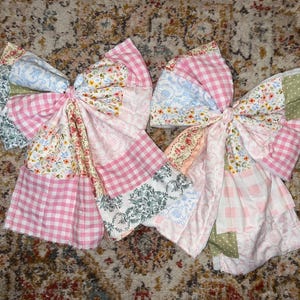 May include: Two large fabric bows with a patchwork design. The bows feature a variety of patterns, including pink gingham, floral prints, and damask. The bows are tied in the center with a matching fabric.