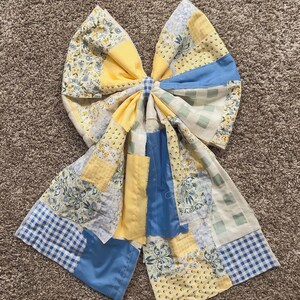 May include: A large, handmade bow featuring a patchwork design. The bow incorporates various fabrics in shades of yellow, blue, and white, with floral and gingham patterns. The bow's center is tied with a matching fabric, creating a decorative accent.
