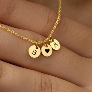 May include: A gold-colored necklace with three small, round pendants. One pendant has the letter "B", another has a black heart, and the third has the letter "A". The necklace is displayed on a person's skin.