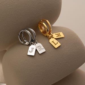 May include: Two pairs of hoop earrings. One pair is silver with two rectangular charms, each with a letter. The other pair is gold with two rectangular charms, each with a letter. The earrings are displayed on a neutral surface.
