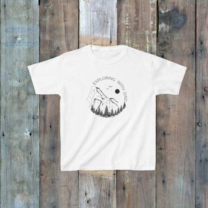 Puede incluir: Camiseta blanca con un gráfico negro de montañas, árboles, un sol y dos pájaros. El texto "EXPLORING WITH DAD" está impreso alrededor del gráfico. La camiseta se muestra sobre un fondo de madera.